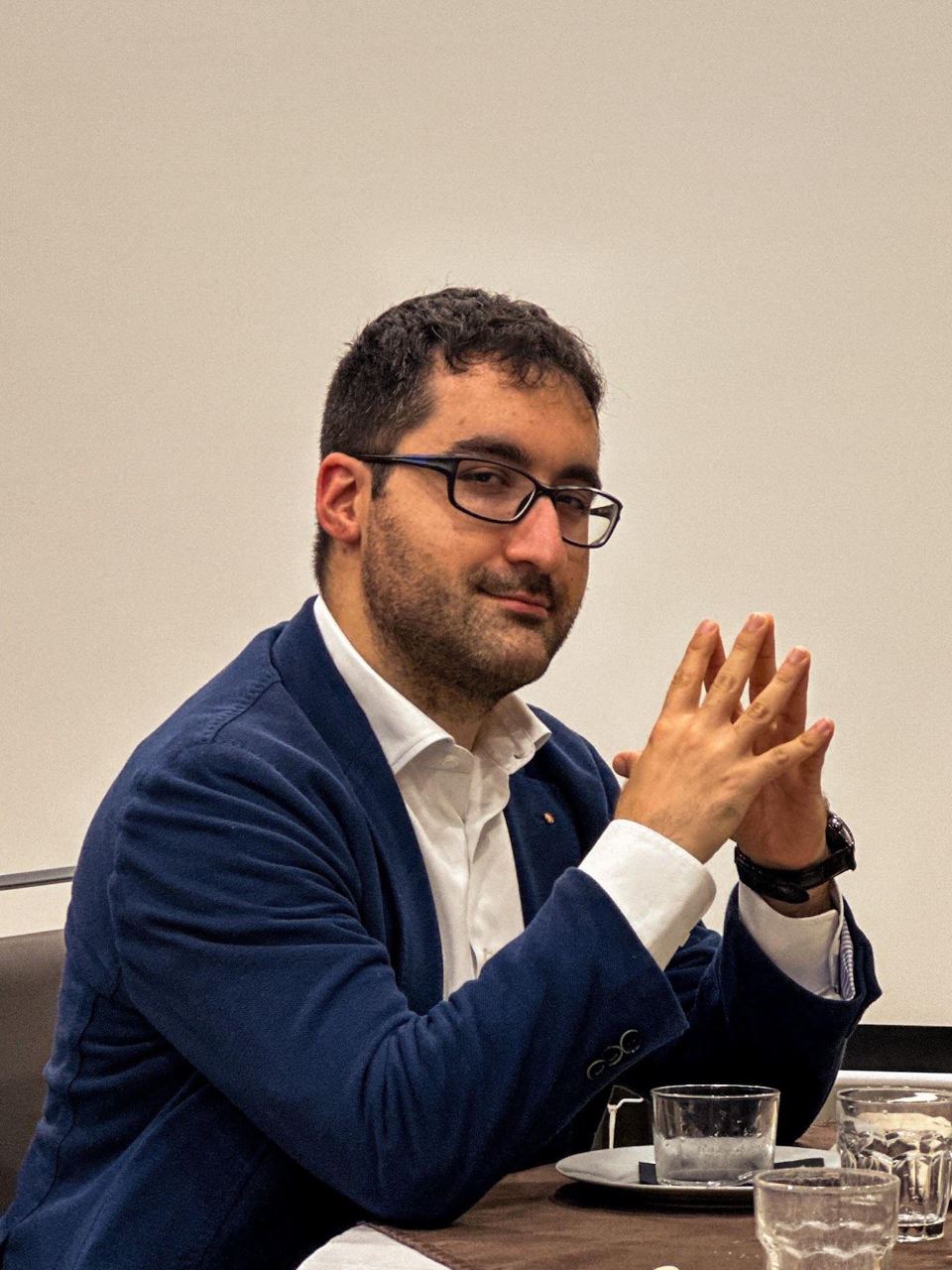 Ritratto: un ragazzo con gli occhiali, seduto a un tavolo con le mani giunte.