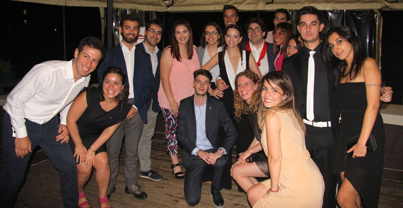 Un folto gruppo di ragazze e ragazzi in abiti eleganti posa sorridente sotto un gazebo, di sera.