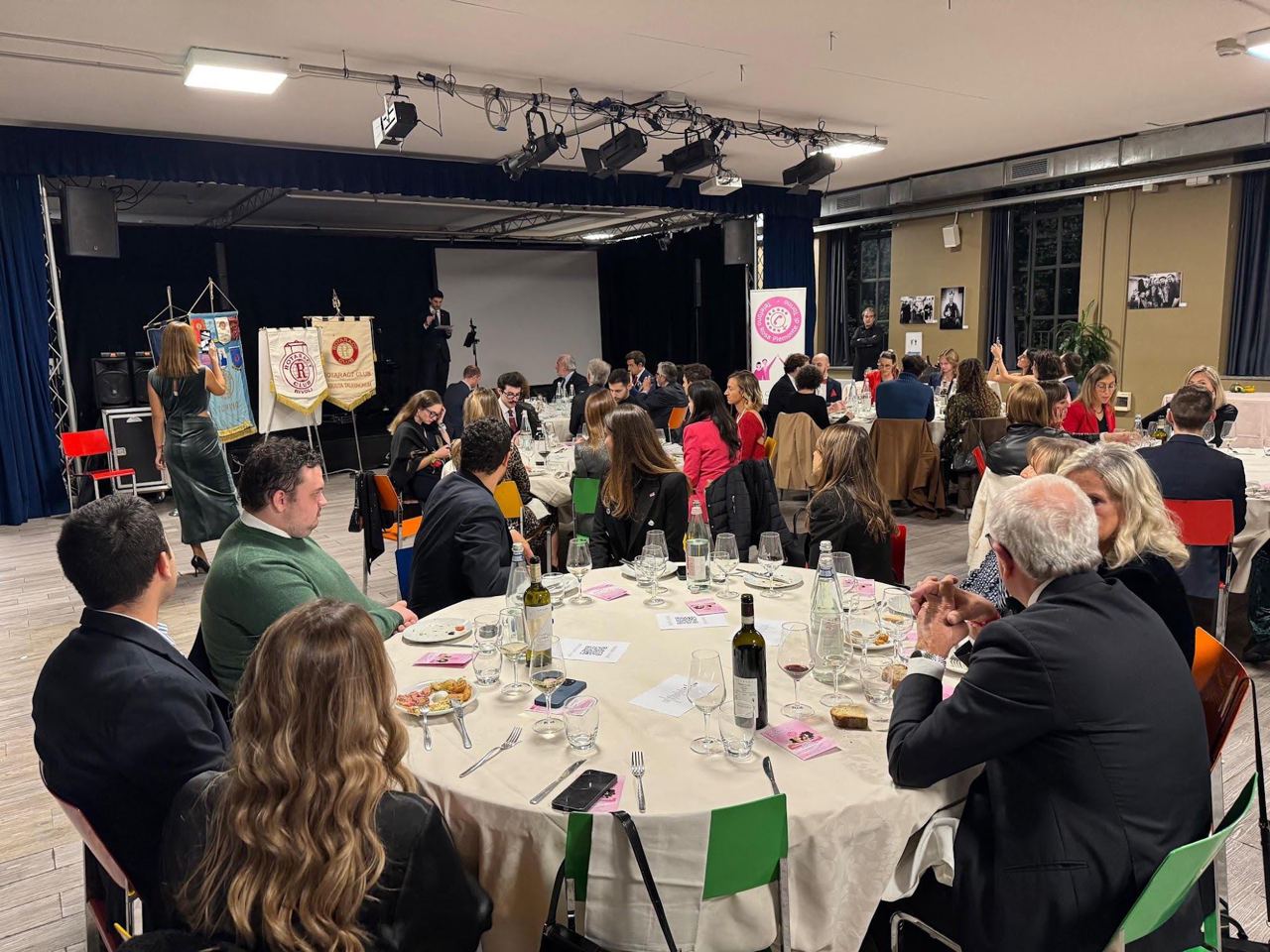 La sala di una cena di beneficenza: tavoli rotondi apparecchiati con tovaglie bianche, ospiti seduti e, sul fondo, gli stendardi dei club.