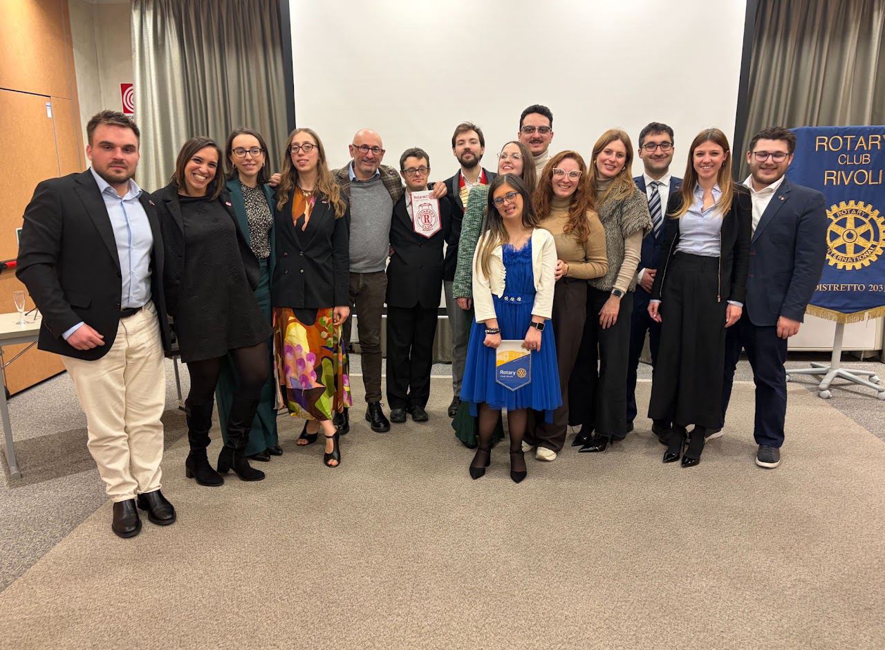Foto di gruppo alla cena con il CEPIM: ragazze, ragazzi e adulti in piedi in una sala, accanto allo stendardo del Rotary Club Rivoli.
