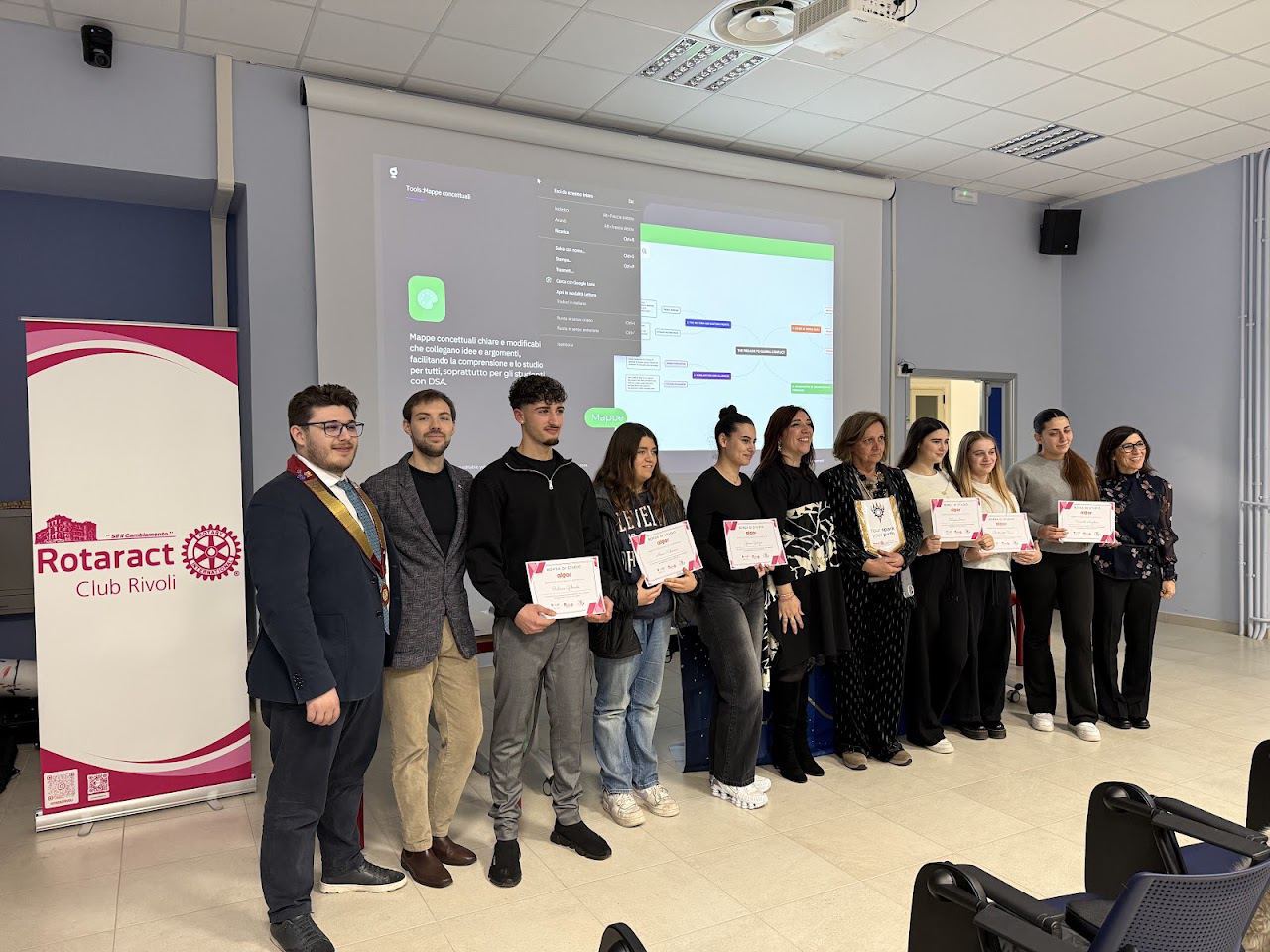 Studenti in fila con gli attestati in mano accanto al roll-up del Rotaract Club Rivoli, in un’aula con un proiettore.