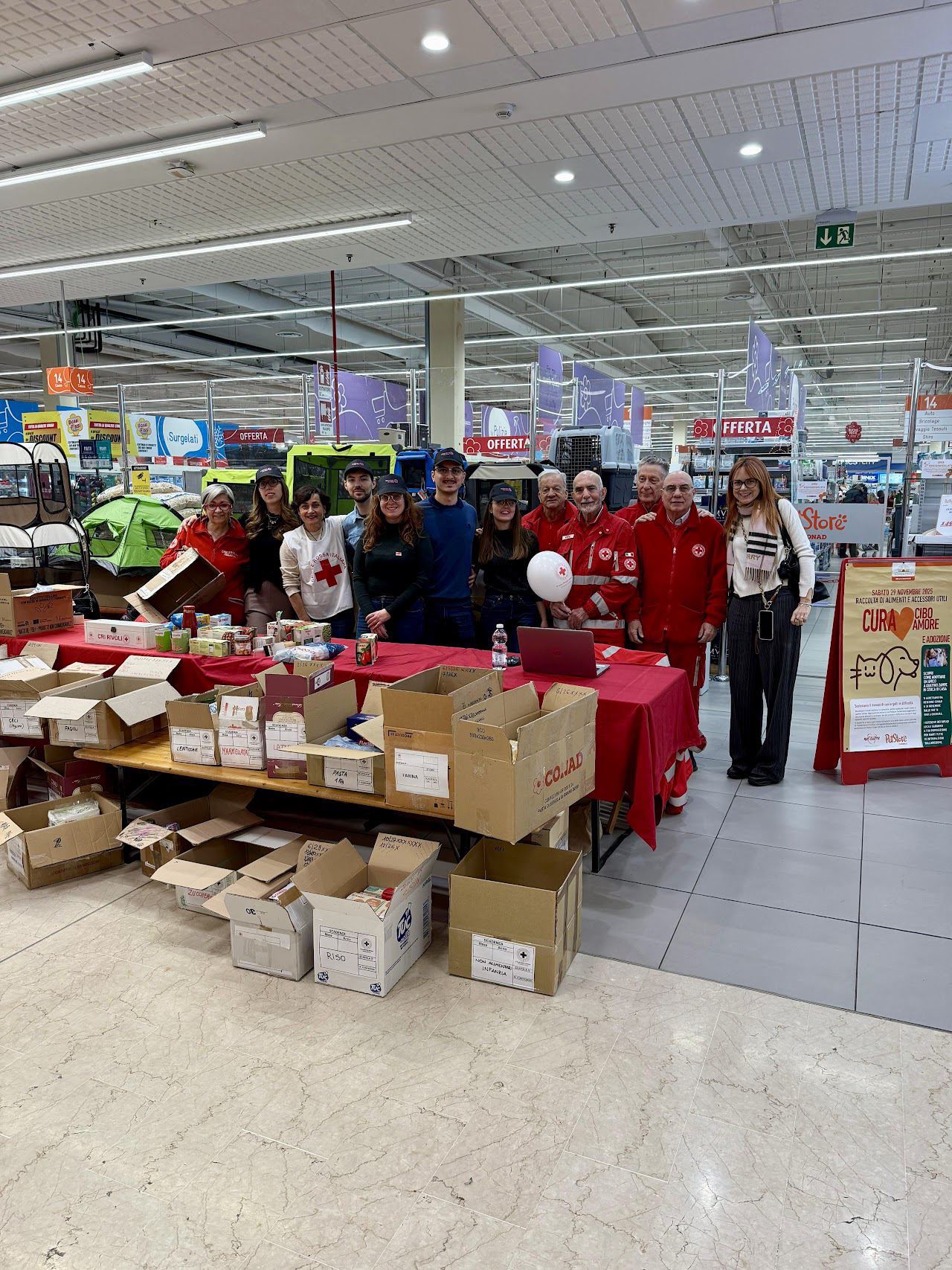 Volontari della Croce Rossa e ragazzi del club dietro un tavolo con gli scatoloni della Colletta Alimentare, in un supermercato.