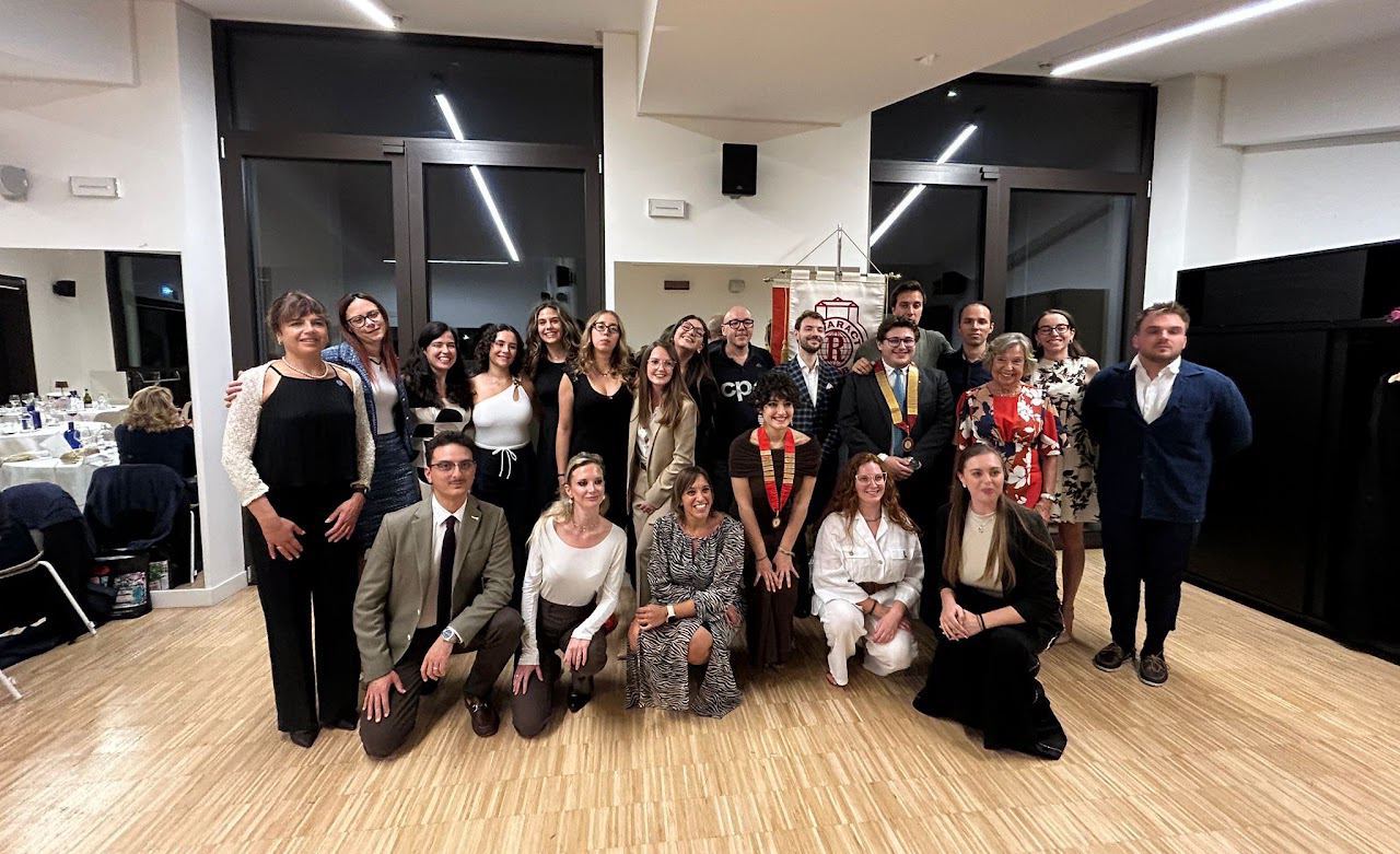 Foto di gruppo al Passaggio di Consegne: ragazze, ragazzi e ospiti in abito elegante in una sala con il pavimento in legno.