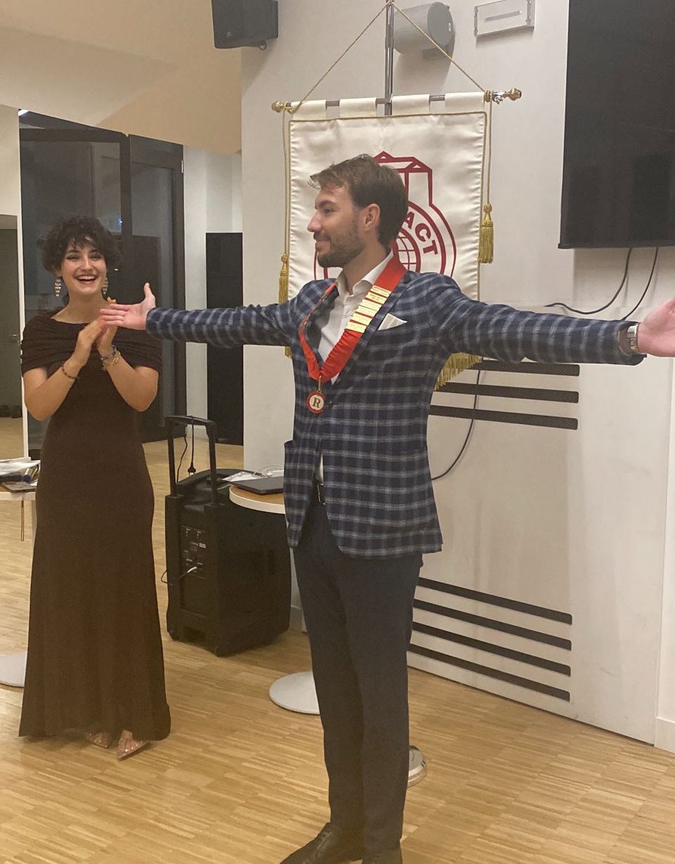 Un ragazzo con il collare del club allarga le braccia accanto a una ragazza che applaude, davanti allo stendardo del Rotaract.