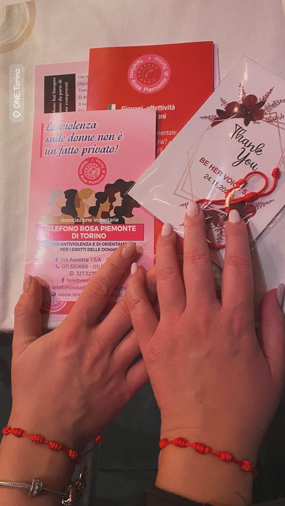 Due mani con il braccialetto rosso, il biglietto di ringraziamento di Be Her Voice e l’opuscolo del Telefono Rosa Piemonte di Torino.