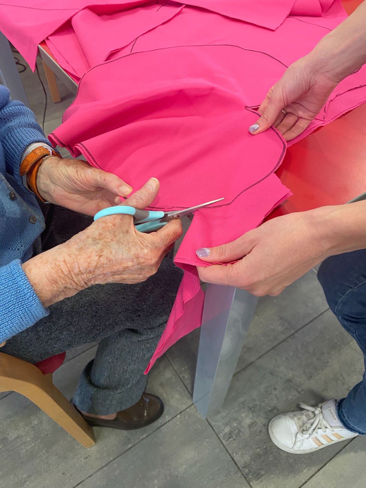 Le mani di un’anziana tagliano con le forbici un tessuto rosa tenuto da mani più giovani