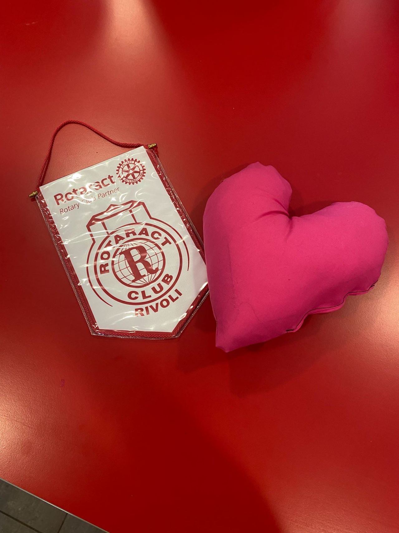 Un cuscino rosa a forma di cuore accanto al gagliardetto del Rotaract Club Rivoli, su un tavolo rosso