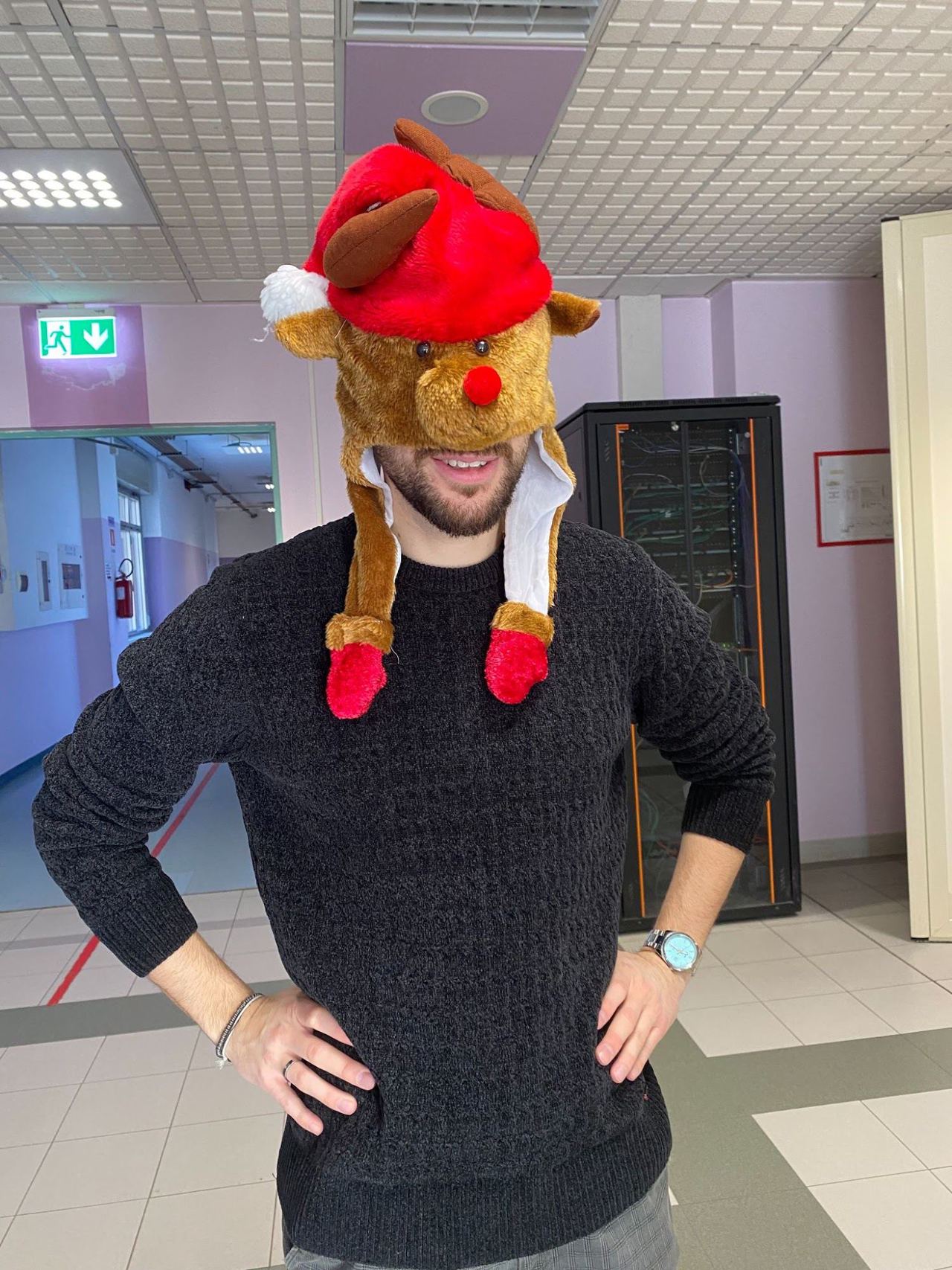 Un ragazzo sorridente, con le mani sui fianchi, indossa un cappello a forma di renna con il berretto di Babbo Natale, in un corridoio.