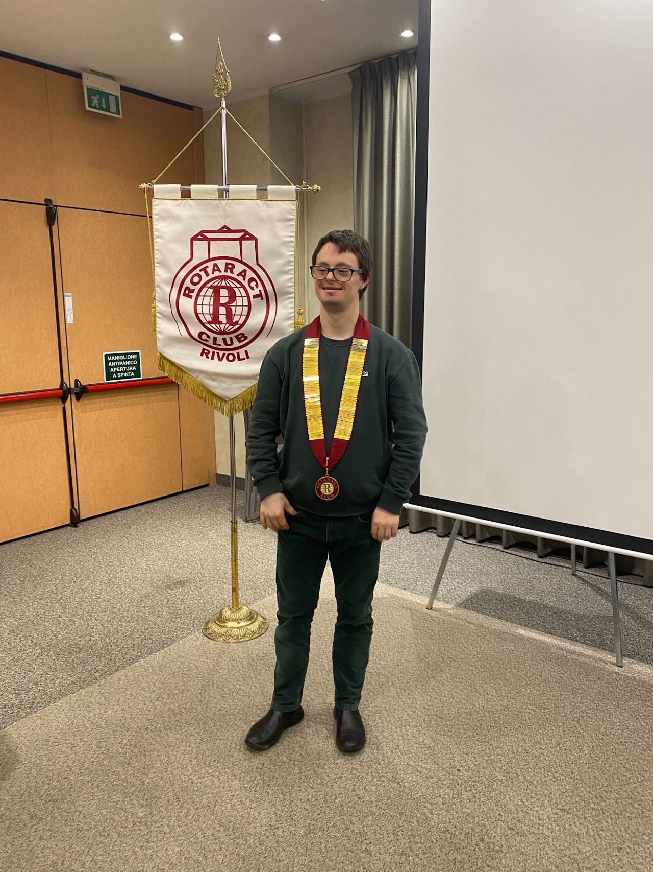 Un ragazzo con il collare del club in posa accanto allo stendardo del Rotaract Club Rivoli.