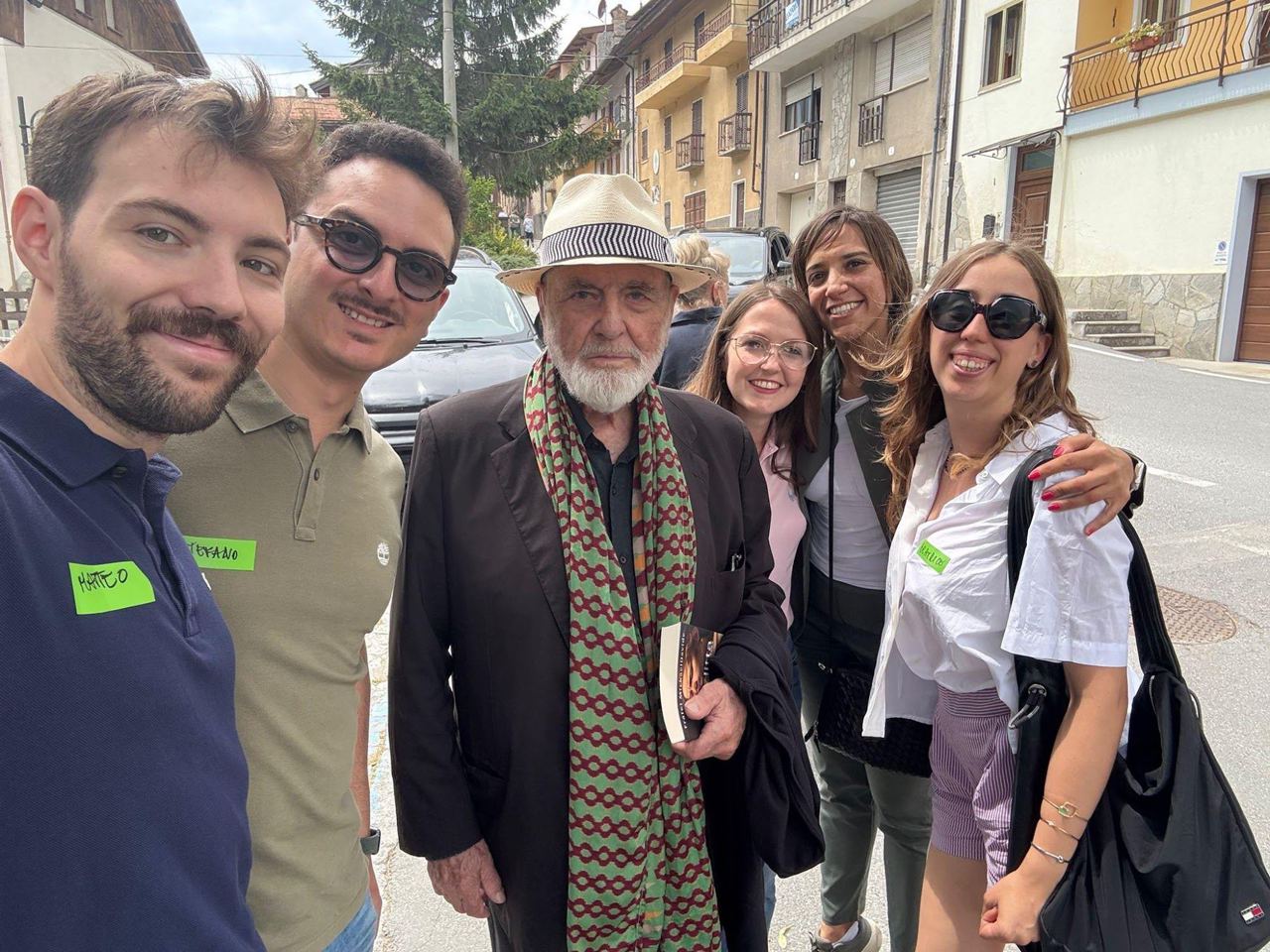 Selfie di gruppo in una via di Exilles: ragazze e ragazzi sorridenti attorno a un signore con cappello di paglia e sciarpa